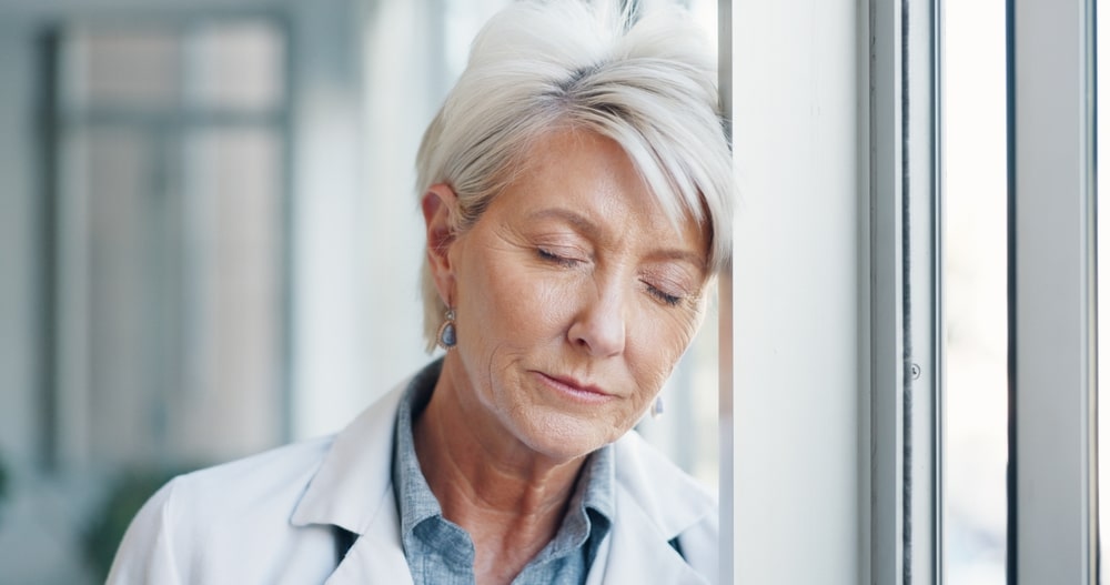 Doctor, sad and burnout of a senior healthcare employee at a window with depression about stress. Health consulting anxiety