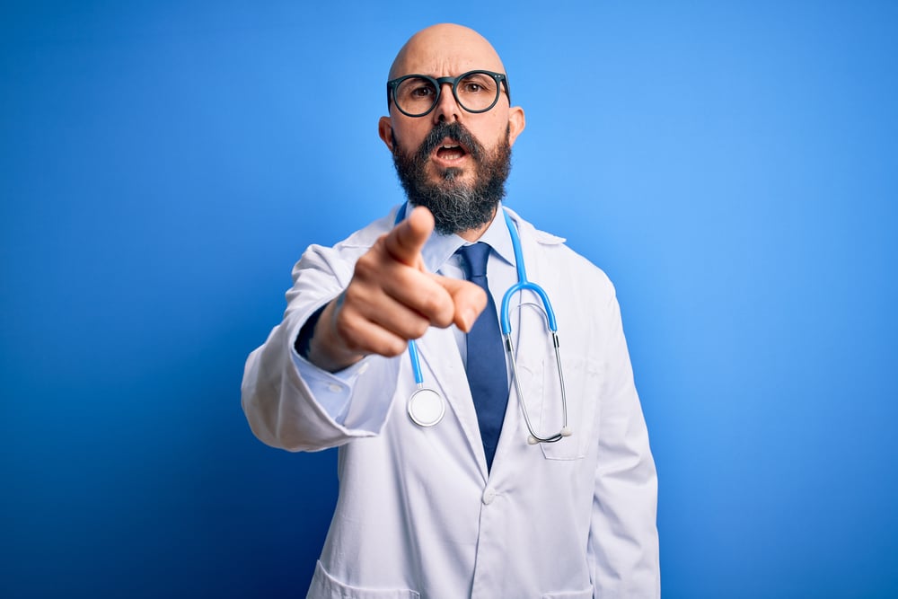 Handsome bald doctor man with beard wearing glasses and stethoscope over blue background pointing displeased and frustrated to the camera