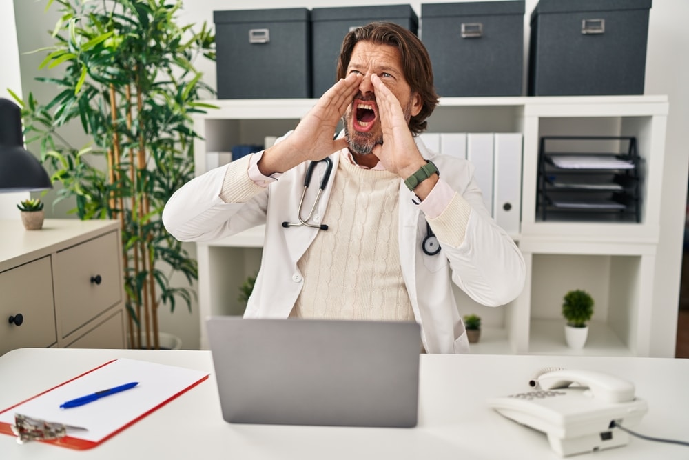 Handsome middle age doctor man working at the clinic shouting angry out loud with hands over mouth