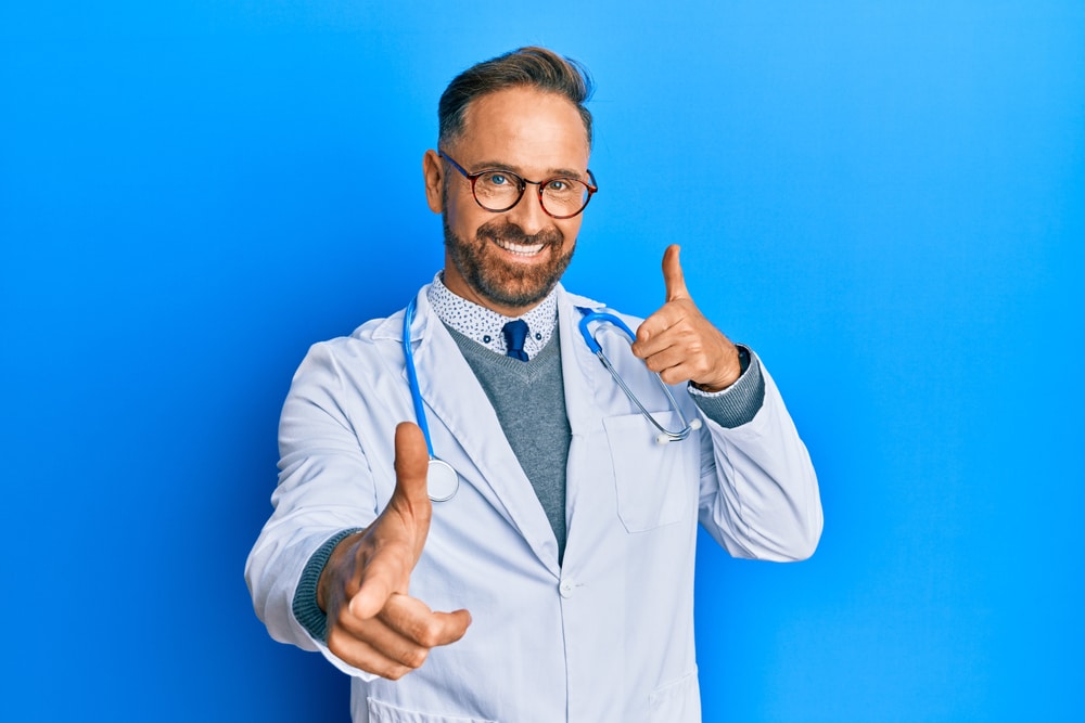 Handsome middle age man wearing doctor uniform and stethoscope pointing fingers to camera with happy and funny face. good energy and vibe