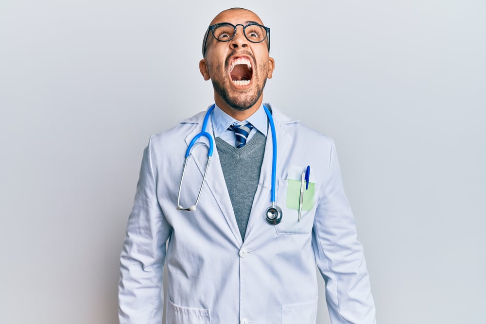 Hispanic adult man wearing doctor uniform and stethoscope angry and mad screaming frustrated and furious, shouting with anger. rage and aggressive