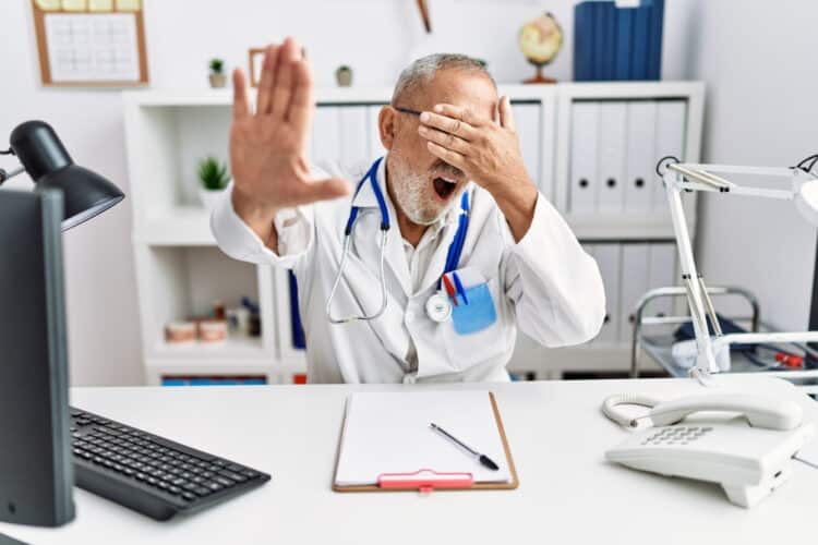 Mature doctor man at the clinic covering eyes with hands and doing stop gesture with sad and fear expression. embarrassed and negative concept