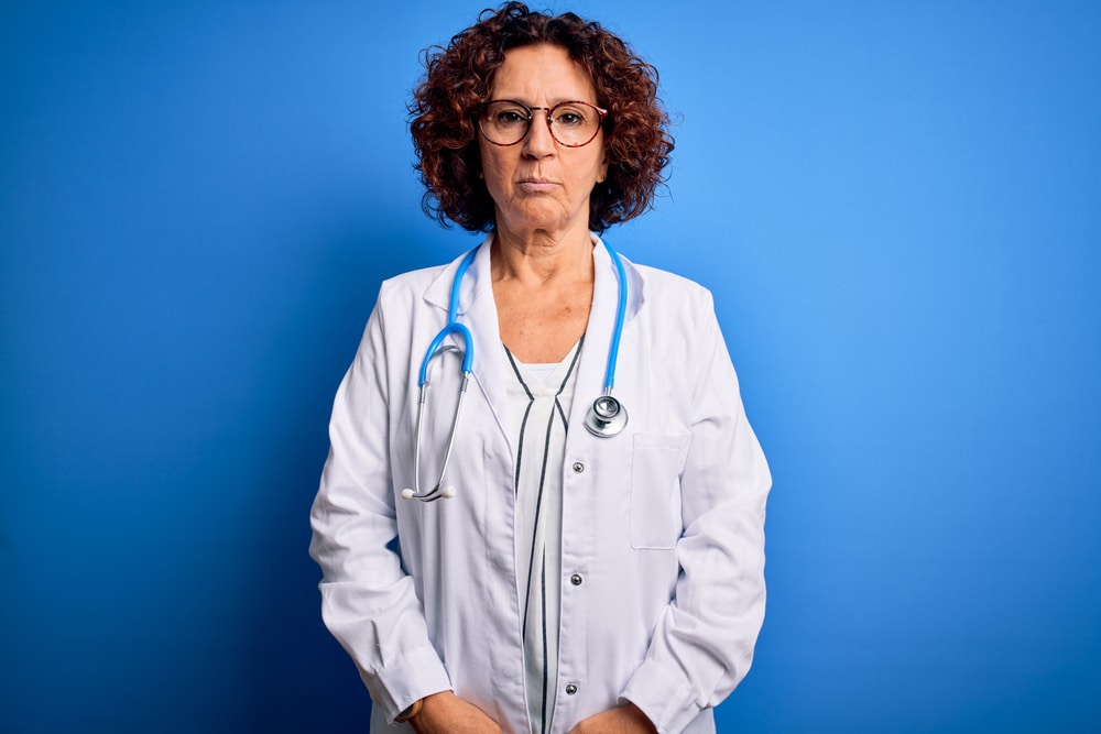 Middle age curly hair doctor woman wearing coat and stethoscope over blue background depressed and worry for distress, crying angry and afraid