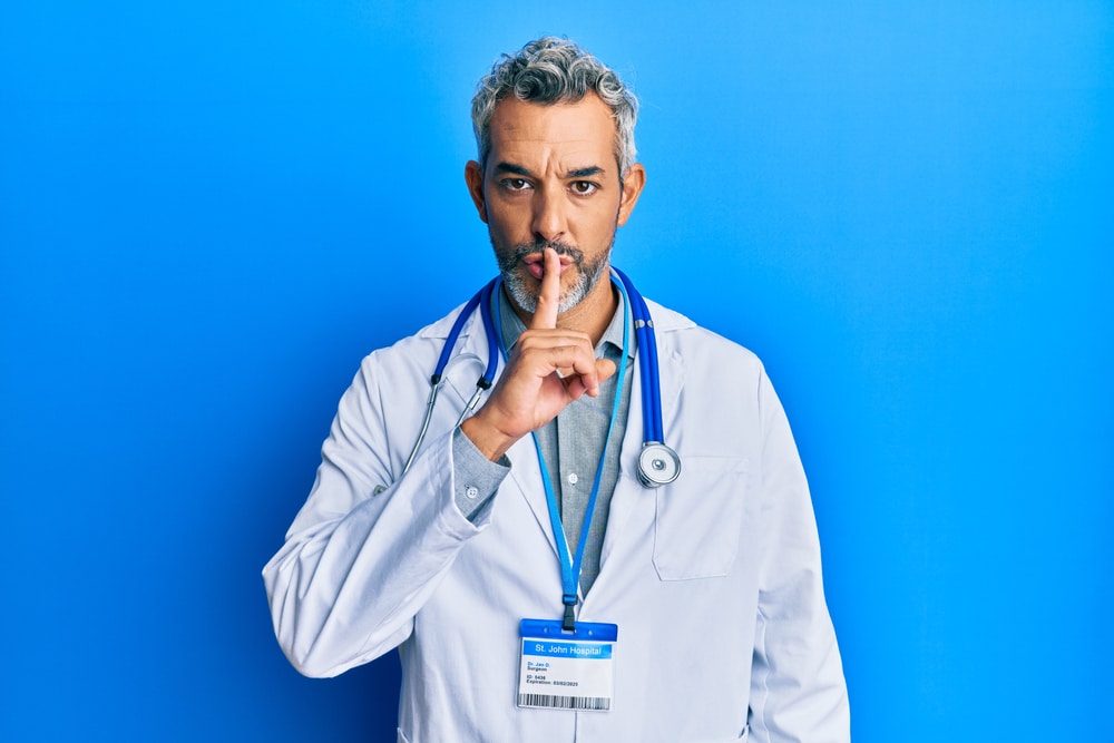 Middle age grey-haired man wearing doctor uniform and stethoscope asking to be quiet with finger on lips. silence and secret concept.