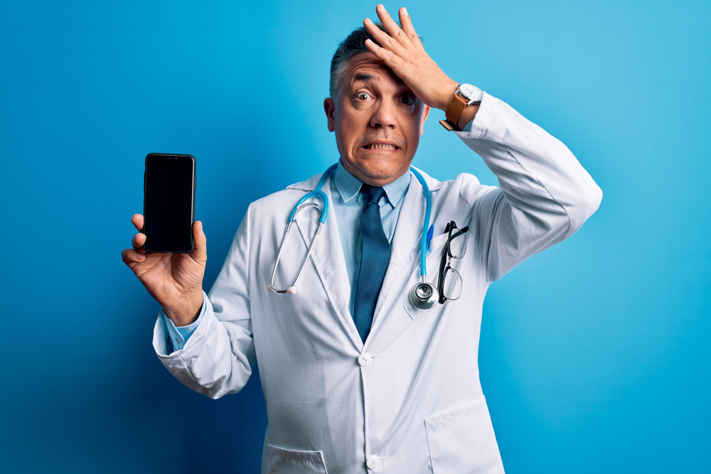 Middle age handsome grey-haired doctor man holding smartphone showing screen stressed with hand on head, shocked with shame and surprise face, angry and frustrated. Fear and upset for mistake