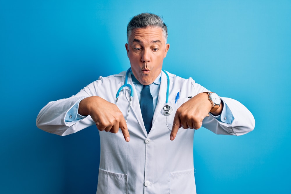 Middle age handsome grey-haired doctor man wearing coat and blue stethoscope Pointing down with fingers showing advertisement, surprised face and open mouth