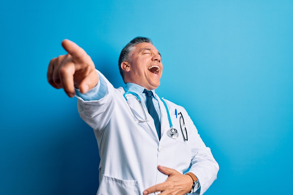 Middle age handsome grey-haired doctor man wearing coat and blue stethoscope laughing at you, pointing finger to the camera with hand over body, shame expression