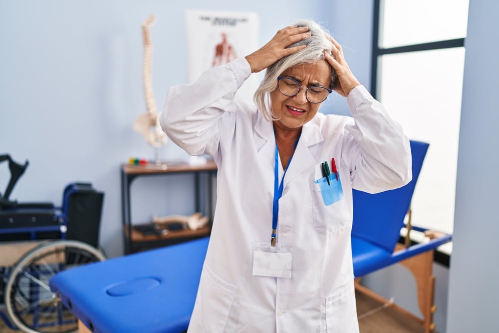 Middle age woman with grey hair working at pain recovery clinic suffering from headache desperate and stressed because pain and migraine. hands on head