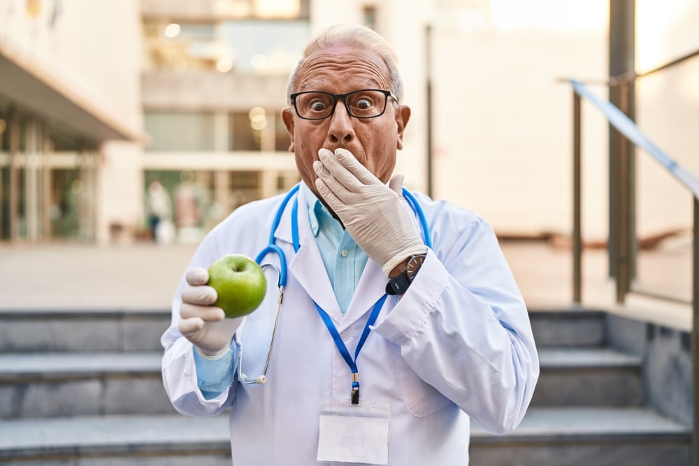 Senior doctor with grey hair holding healthy green apple covering mouth with hand, shocked and afraid for mistake. surprised expression