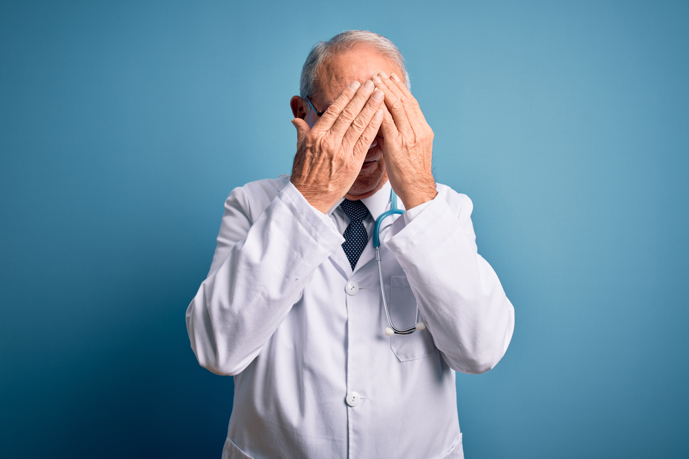 Senior grey haired doctor man wearing stethoscope and medical coat over blue background with sad expression covering face with hands while crying