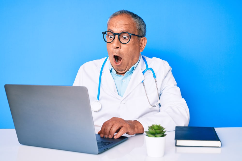 Senior handsome man with gray hair wearing doctor uniform working using computer laptop afraid and shocked with surprise expression, fear and excited face