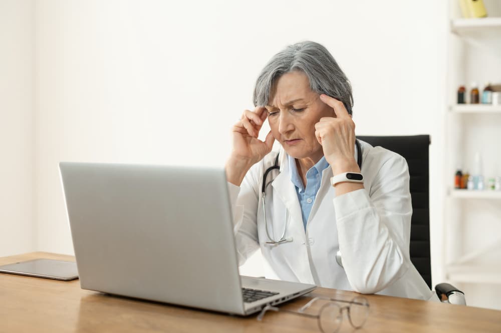 Tired depressed senior female doctor sitting alone in the office in front of a laptop, feeling burnout at work or desperate and thinking of a medical problem