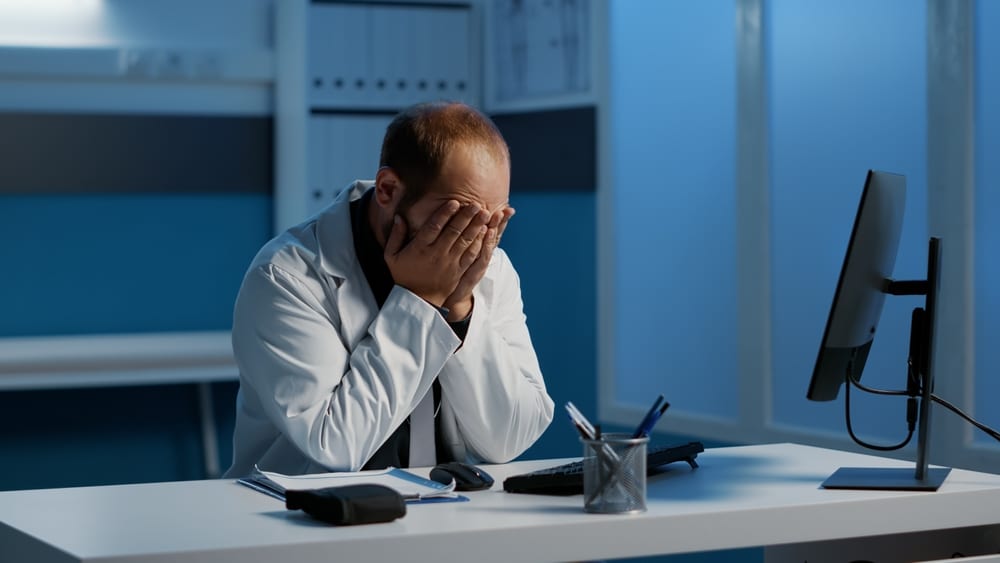 Tired exhausted physician man working night shift in hospital office analyzing medical expertise on computer typing health care report. Stressed doctor planning planning patient treatment