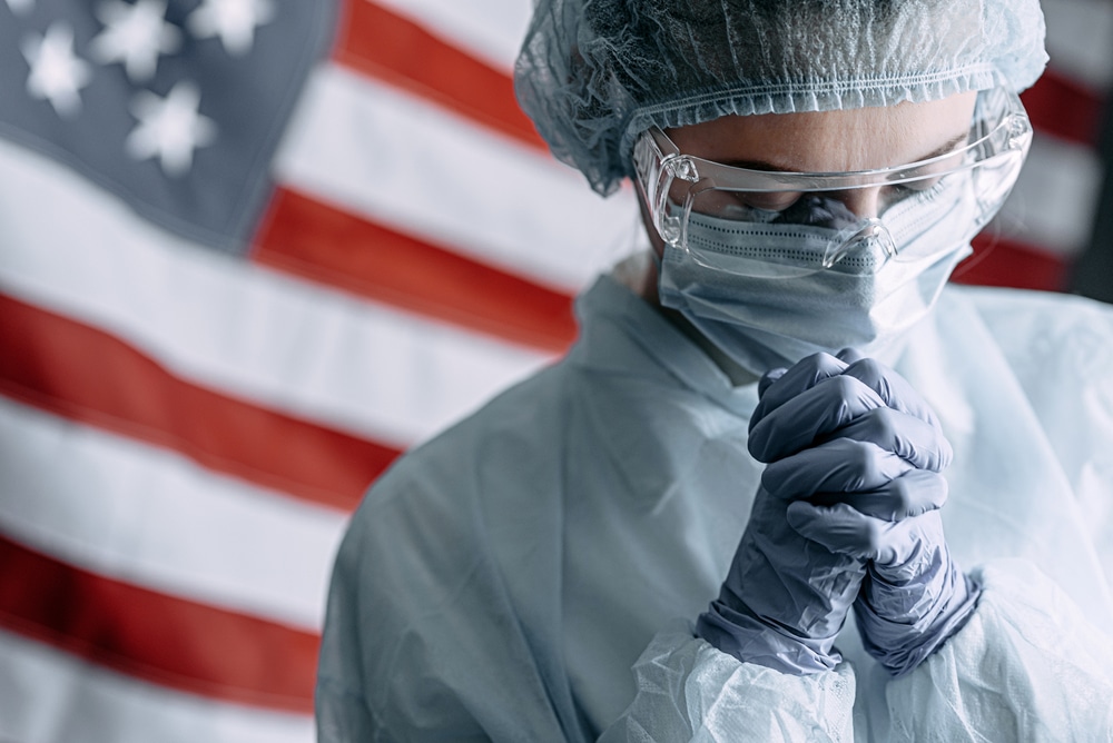 young nurse in a protective medical mask and hat and dressing gown prays against the backdrop of the American flag and asks God for help and strength in the fight against the coronavirus epidemic