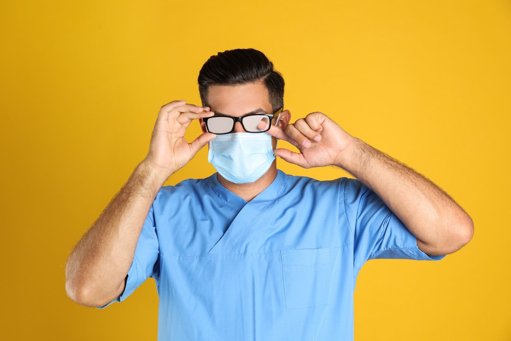 Doctor wiping foggy glasses caused by wearing medical mask on yellow background