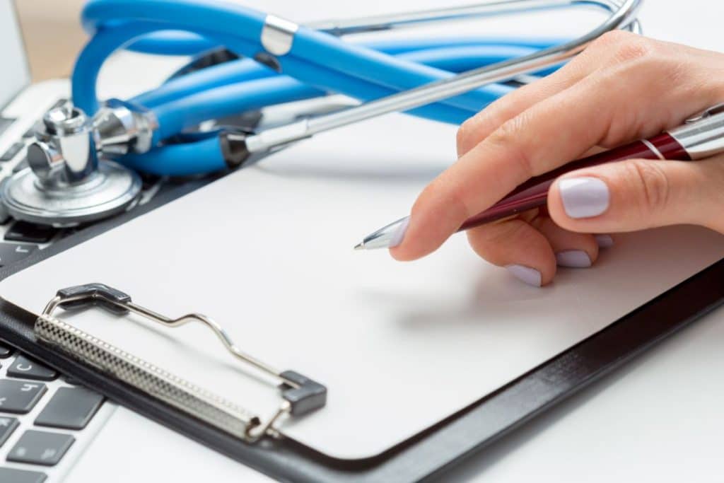 medical scribing, hand holding a pen over the clipboard with stethoscope and laptop