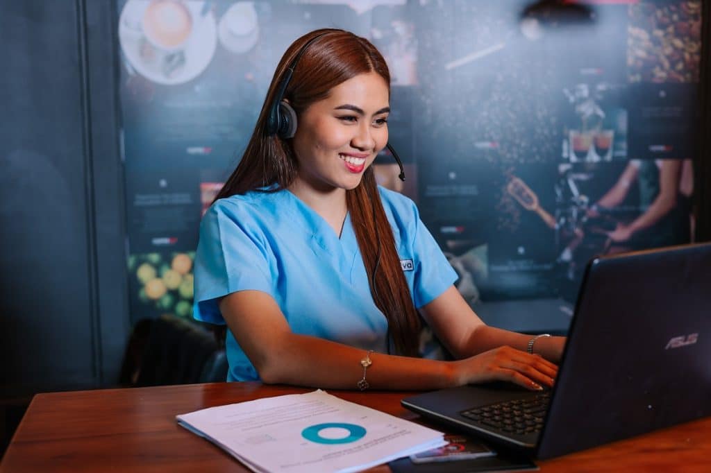 A female ER Scribe in blue scrubs and a headset is smiling while typing on a laptop