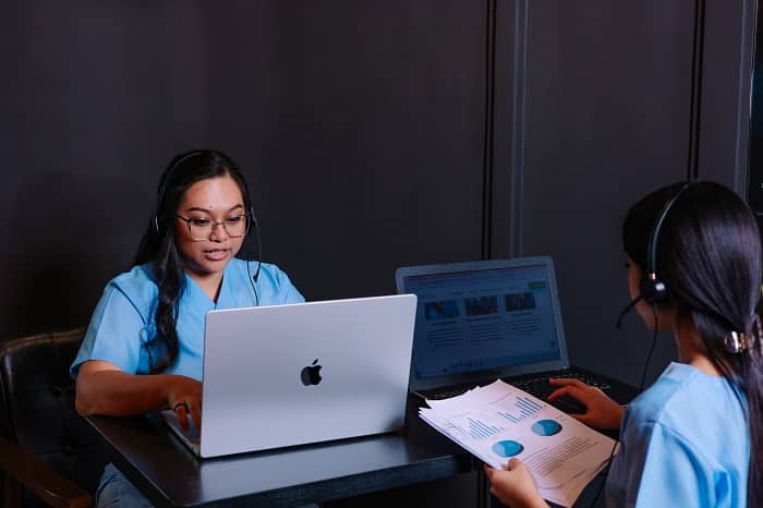 A medical biller is holding a document with charts while talking to a colleague working on a laptop.