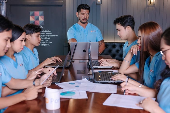Cardiology billing services team in blue scrubs working on laptops at a dark wooden conference table.