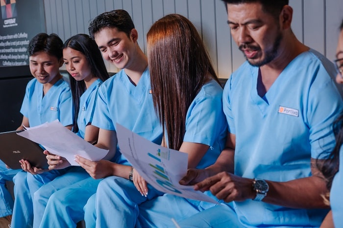 Five medical professionals in blue scrubs sit in a row, reviewing documents and smiling.