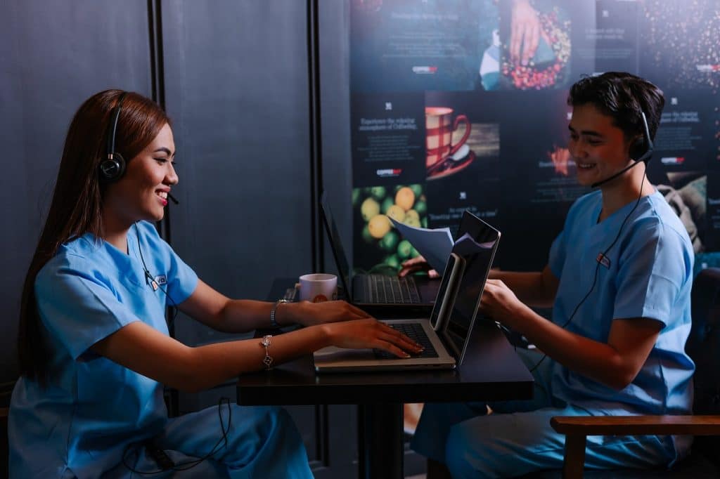 Two ER Scribe professionals in medical scrubs working on laptops with headsets, documenting patient information in a hospital setting