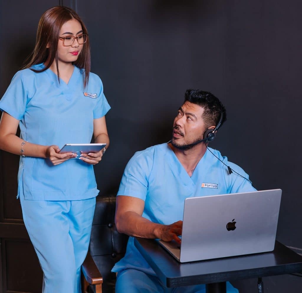Two healthcare professionals in blue scrubs collaborate using a laptop and tablet, representing revenue cycle management services for small practices in a modern clinical office setting.