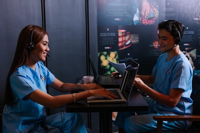 Two smiling colleagues in scrubs working on laptops.