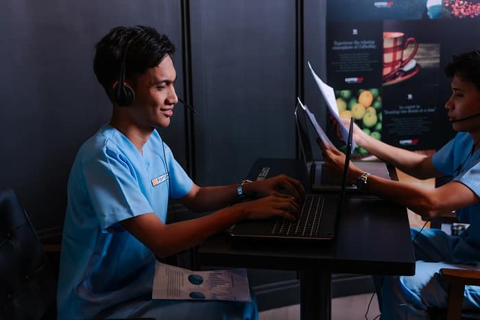 Two smiling men in blue scrubs and headsets work on laptops and review financial reports at a dark table.