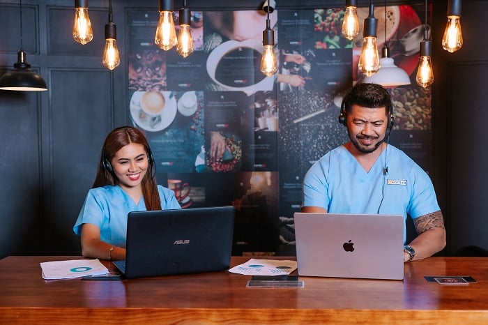 Two smiling professionals in light blue scrubs and headsets work on laptops at a wooden counter.