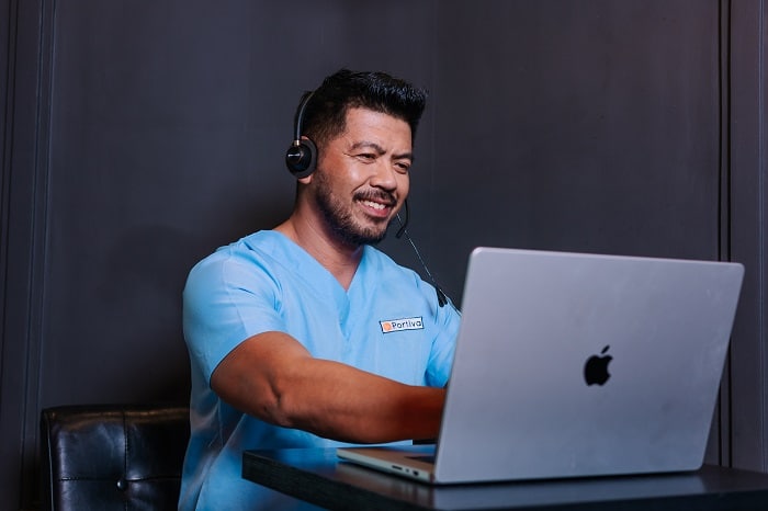 A man in a light blue scrub top is wearing a headset and smiling while working on a laptop.