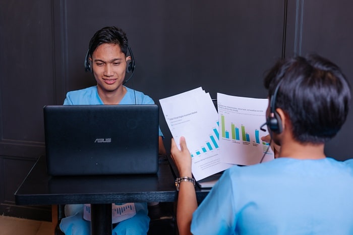 two individuals in blue scrubs and headsets work at desks, one on a laptop, the other reviewing charts