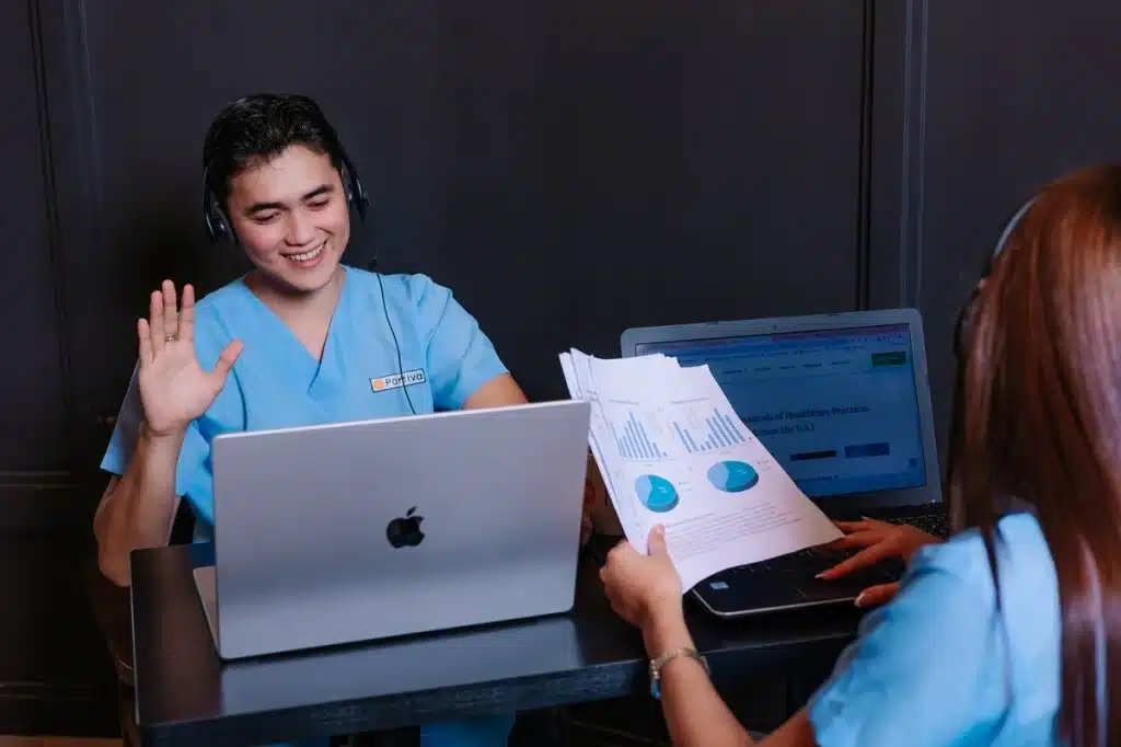 AI patient communication workflow specialist wearing a headset smiles and waves during a virtual meeting while working on a laptop