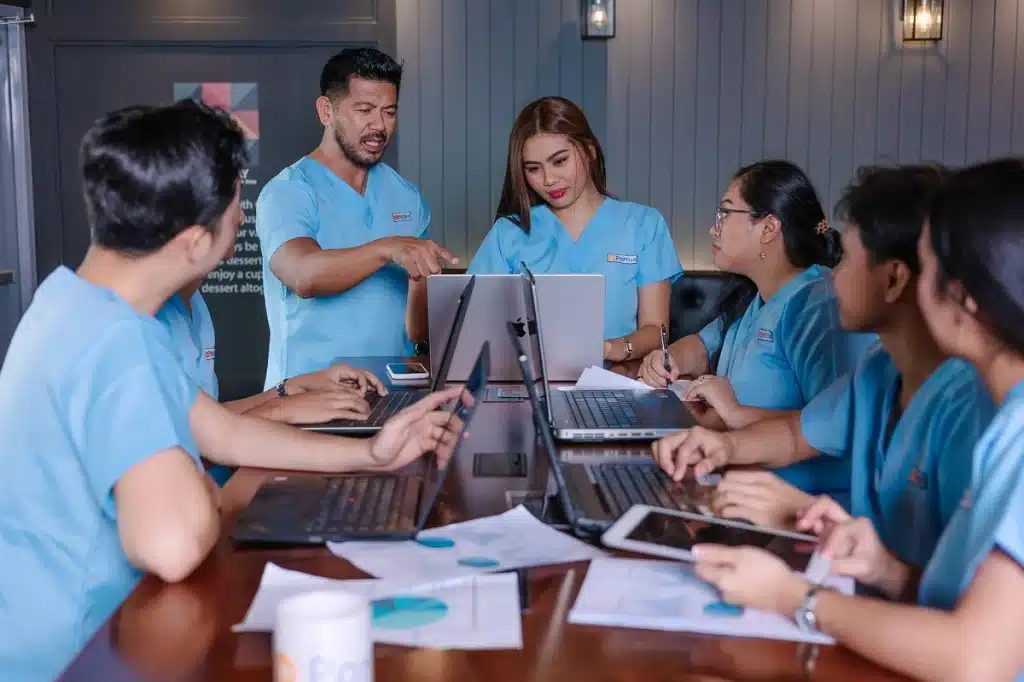 Medical receptionist scheduling workflow in action as receptionists in blue scrubs collaborate over laptops and tablets to review patient appointments, follow-ups, and workflow processes